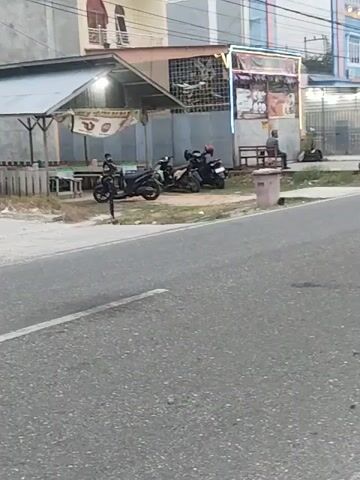 The video captures a street scene. A white car is driving on the road. To the left of the road, there are several parked motorcycles. In front of the motorcycles is a small covered structure with tables and chairs. Behind the motorcycles, there is a building with windows and some decorative lights. To the right of the motorcycles, a person is sitting on a bench. The scene appears to be in a built-up area, possibly a commercial or residential district. The overall lighting suggests it is either early morning or late afternoon.