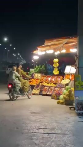 The video captures a lively nighttime scene at a roadside fruit stand in Kandahar, Afghanistan. The dark sky is illuminated by numerous streetlights that line a bustling road stretching into the distance. The fruit stand is brightly lit by several overhead bulbs, showcasing a vibrant array of fresh produce.

Various fruits are neatly arranged in crates and baskets, including large piles of oranges, watermelons, bananas hanging from above, grapes, apples, and pomegranates. A fruit vendor, dressed in traditional clothing, stands behind the display, attending to customers.

The central activity shows a motorcycle approaching the stand. An adult man is driving, with a child seated in front of him. They stop briefly at the fruit stand, and the man interacts with the vendor, who hands him a white plastic bag, indicating a purchase. After the quick transaction, the motorcycle drives off. Another person can be seen standing near the fruit display. The scene depicts a common evening market interaction, characterized by the glow of artificial lights against the dark backdrop of the night. No visible text is present in the frame.