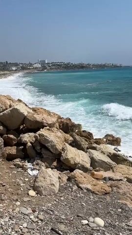 The video shows a rocky shoreline with turquoise ocean waves crashing against the rocks. In the background, a distant coastal town with buildings is visible under a clear blue sky. The camera is positioned at ground level, looking out towards the sea, with a close-up view of the rocks in the foreground. There are no people or animals visible in the frame. The environment appears to be a sunny day with moderate waves.