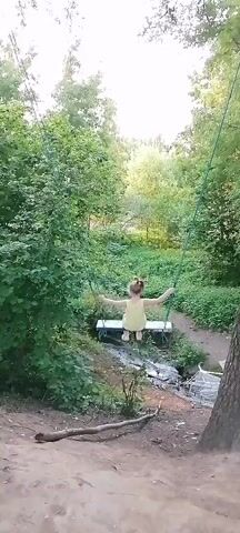 A child in a yellow dress is swinging on a swing in a wooded area. The child is swinging back and forth with their arms outstretched. The swing is made of a wooden seat and ropes attached to a tree branch. The background is filled with trees and greenery. The sun is shining through the trees, creating a dappled light effect.