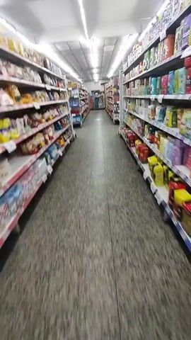 The camera moves through a grocery store aisle, looking straight ahead. On either side are tall shelves stocked with food items including snacks, cookies, canned goods, and baking supplies. The floor is gray with a speckled pattern. The ceiling is white with rows of fluorescent lights.