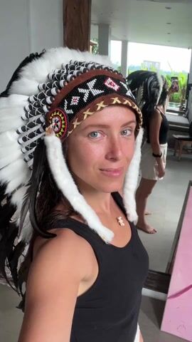 A woman wearing a Native American headdress is standing indoors, looking at her reflection in a mirror. She is wearing a black tank top and white shorts. The headdress is primarily white with black accents and intricate beadwork. Behind her, another person is also wearing a similar headdress and holding a phone, possibly taking a picture. The interior setting appears to be a well-lit room with a view to the outside. The environment suggests a casual or touristy atmosphere.