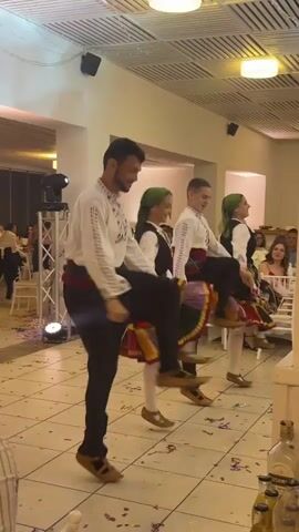 Four people dressed in traditional clothing are dancing in a banquet hall. Two men are wearing white shirts embroidered with red and black designs, black pants, and traditional sandals. The two women are wearing dark dresses with embroidered skirts, headscarves, and similar sandals. They are dancing in unison, moving in a line along the tiled floor, with tables and seated guests visible in the background. Confetti litters the floor. A stage lighting fixture stands to the left, and bottles of Ushmi drink stand in the lower right corner. The room is lit by overhead fluorescent panels.