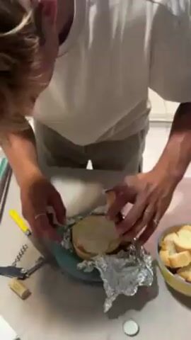 A man with blond hair, wearing a white t-shirt and khaki shorts, cuts into a wheel of brie cheese on a light blue plate. The cheese is wrapped in aluminum foil. A yellow-handled knife and bottle opener are to the left of the plate, while a yellow bowl full of sliced bread sits to the right. The man wears rings on both ring fingers.  The scene takes place on a light-colored countertop in a kitchen setting. Electronic dance music is playing in the background.