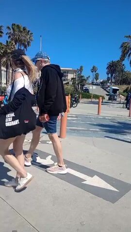 The video captures a sunny day at a beach parking area, where cyclists are riding and passing. Several people on bikes pass by the camera's field of view, heading in the direction indicated by a large arrow painted on the ground. Palm trees line the area alongside the parking lot, and in the background, there's an incline leading to a street lined with cars and a couple of buildings. The camera remains stationary throughout the short video.