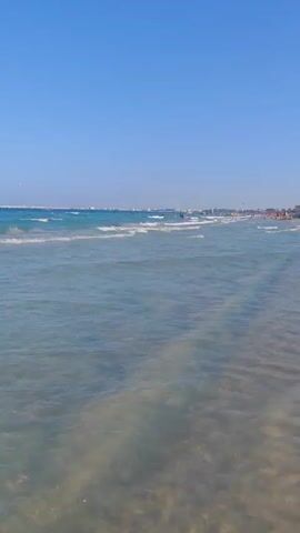 The video shows a beach with clear blue sky and calm ocean waves. The water is shallow near the shore, allowing the sandy bottom to be visible. Several people are in the water, some swimming and others standing. The waves are small and gently break as they approach the shore. The sun is shining, creating a bright and warm atmosphere. There is no discernible movement in the foreground or midground, other than the natural movement of the water. The scene is peaceful and idyllic, conveying a sense of relaxation and enjoyment.