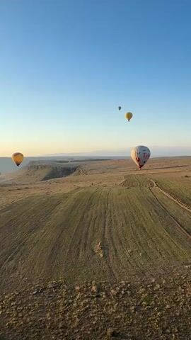 The video shows a serene aerial view of multiple hot air balloons floating over a vast, arid landscape at sunrise or sunset. The sky transitions from a clear blue overhead to a warm yellow and orange glow near the horizon. Below, the terrain consists of dry, brown earth interspersed with patches of green, possibly cultivated fields, showing distinct parallel lines. In the distance, flat-topped hills and mountains create a rugged backdrop.

Several hot air balloons are visible:
- A prominent white balloon with red lettering (partially visible, suggesting "Tim Hortons") is seen on the right side of the frame initially.
- A yellow and orange balloon is on the left side, gradually moving towards the center as the camera pans.
- Two smaller, yellow balloons are higher up in the distance.
- Another, more distant, lighter-colored balloon is also visible.

The camera, briefly identifiable as a phone on a stick at the top left of the frame, pans slowly from right to left, revealing more of the expansive landscape and the scattered balloons. The overall scene suggests a peaceful and picturesque hot air balloon experience in the region of Yeşilhisar, Turkey.