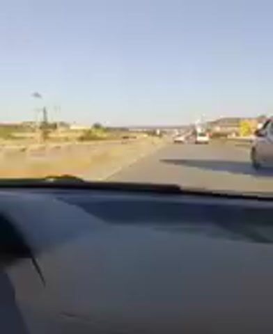 The video is shot from the perspective of someone in a car, looking forward. The car is driving on a highway. There are other cars and trucks on the road. On the sides of the highway, there are signs and vegetation. In the distance, there are buildings and hills. The sky is clear and blue. The video captures a typical highway driving scene with various vehicles and roadside elements.