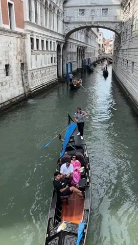 A gondola navigates a Venetian canal. The gondola holds four people including the gondolier who stands at the rear, propelling the boat with a long oar. The other three passengers sit in the gondola’s seating area. The canal is lined with tall, ornate buildings of light-colored stone and brick, some of which are connected by arched bridges overhead. Several other gondolas are visible in the distance. The sky is overcast.
