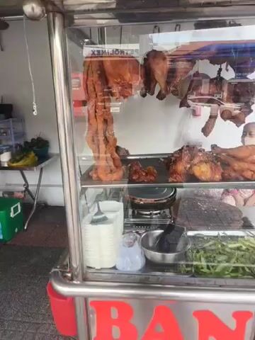 The video shows a close-up view of a glass display case filled with roasted meats, primarily poultry. The meats are hanging and displayed on racks. A young woman with dark hair pulled back is visible behind the display case, looking towards the camera. The background appears to be an outdoor setting with some structures and a hint of greenery. The lighting is bright, suggesting daytime. The overall impression is of a food vendor stall or a market stall specializing in roasted meats.