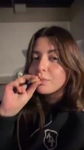 A young woman with shoulder-length brown hair and a nose ring is seen holding a lit cigarette between her lips and her fingers.  She has a necklace with a small shield pendant. The background shows a white wall and part of what appears to be a kitchen area. She exhales smoke towards the camera.