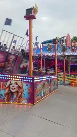 The video shows a carnival ride with several people on it. The ride is a pendulum-style attraction that swings back and forth. The people on the ride appear to be enjoying themselves, with some of them smiling and laughing. The ride is decorated with colorful lights and images, and there are flags on poles around the ride. The sky is cloudy, suggesting it may be an overcast day. In the foreground, there is a pink platform with an image of a woman on it and the words "SING TIME" and "SWING."