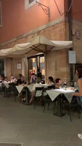 The video captures a charming evening scene in what appears to be a European town. The setting is a narrow cobblestone street lined with buildings featuring a warm, terracotta-colored facade on the right. Several outdoor dining tables, covered with white tablecloths, are arranged along the sidewalk, creating a cozy restaurant atmosphere.

Several people are seated at these tables, engaged in conversation and enjoying their meals. They appear relaxed and happy, suggesting a pleasant social gathering. A few individuals are also seen walking along the street. One man in a white t-shirt and shorts is walking towards the camera with his arms outstretched, seemingly in a jovial mood. Another person in a white shirt and patterned pants walks away from the camera in the foreground.

The time of day is evident as dusk or early evening, with the sky a deep blue, and artificial lights illuminating the street and restaurant areas. Flags with a red and white checkered pattern are strung across the street, adding a festive touch. The architecture of the buildings, particularly the arched windows and balconies, suggests an old-world European location. A sign is partially visible on the building, though the text is not legible. The overall ambiance is one of a peaceful, communal evening in a picturesque town.