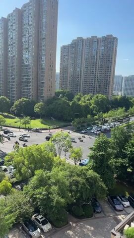The camera pans down to a view of a street and a parking lot from an elevated position. The street has multiple lanes with sparse traffic, including cars, scooters, and what appears to be a bus. Trees line the street and separate the traffic from sidewalks. Pedestrians cross at intersections. In the foreground, a parking lot with multiple parked cars is visible, including a blue and a red car, along with others in neutral colors. High-rise apartment buildings dominate the background. The sky is clear and blue, suggesting a sunny day.