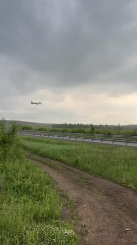 A red and white airplane descends toward the ground on a cloudy day. The plane is seen from a distance, flying over a highway and a grassy field. The highway has a metal guardrail. There are trees and shrubs in the distance. The camera remains static throughout the video.
