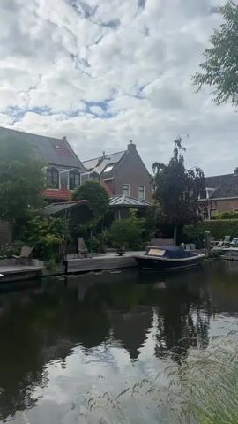 The video shows a canal with houses and boats along the bank under a cloudy sky. The houses are of varying sizes and architectural styles, with some featuring red tile roofs and solar panels. Lush green trees and vegetation surround the properties. A dark-colored boat with a blue cover is docked in the canal, reflecting in the water. The water itself is dark and reflects the sky and the surrounding environment. The foreground shows tall, dry grass. The scene appears to be a residential area with a waterway, possibly in the Netherlands or a similar European country. The overall atmosphere is calm and serene.