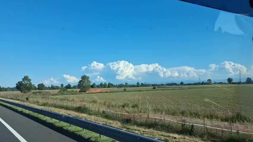 The camera looks out from a car window on a sunny day. A highway is on the left, with a guardrail in the foreground. Beyond the highway is a field of dry grass and crops, bordered by trees. In the distance, there are very large, puffy white cumulus clouds against a bright blue sky.  The camera moves, so the perspective changes.