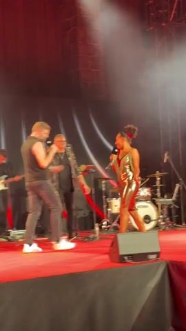 The video shows a live music performance on a stage.  In the foreground, a man and a woman are singing into microphones, appearing to interact with each other. The woman is wearing a shiny, form-fitting dress and high heels, while the man is wearing a black vest over a t-shirt and dark pants.  Behind them, a band is visible, including a guitarist on the left, a keyboardist or another singer in the center, and a drummer on the right. The stage is covered in a red carpet and is lit by spotlights. The background features a dark curtain with red accents and a brick-like structure at the top.