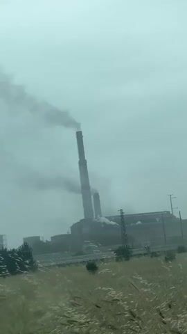 The video shows a factory with smokestacks emitting dark smoke. In the foreground, there is tall grass swaying in the wind. The sky is overcast, and the overall atmosphere is hazy. The factory appears to be industrial, with large buildings and structures visible. There are no people or animals visible in the video. The footage is shot from a distance, with the factory being the main focal point. The video is composed of several short clips, with slight variations in the camera angle and composition.
