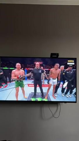 The video shows a television screen displaying a UFC fight. In the first frame, two fighters stand center, a referee is between them, and several people stand at the edges of the ring. The referee raises the arm of one fighter. The second frame shows the fighter with his arms raised, smiling and yelling. The fighter has glasses and multiple tattoos on his chest. A man in a suit can be seen on the right of the screen.