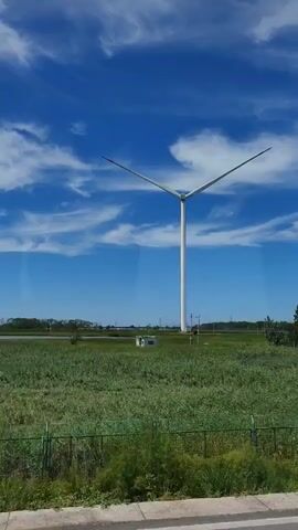 A white wind turbine stands tall against a bright blue sky with wispy white clouds. The turbine has three blades, and two are angled slightly upwards and to the left, while the third is angled downwards and to the right. In the foreground, there is a field of green grass and some vegetation. A white vehicle with a red ribbon on its antenna drives by in front of the wind turbine. Further in the distance, there are some power lines and a small white building. The video pans slightly to the left, revealing a large white tanker truck with Chinese writing on its side. The truck is parked on a road, and the wind turbine is visible behind it.
