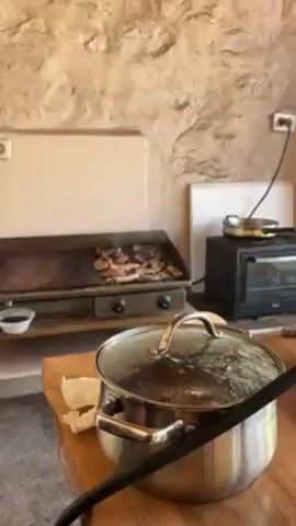 The camera pans up from a covered pot to a flat-top grill with pieces of various meats cooking. The grill is stainless steel with two black knobs on the front and green lights illuminating on either side. Some of the meat appears to be strips of bacon, some chunks of chicken, and some cuts of beef. Grease spatters as the meat cooks.  The background is a wall made of stone and white plaster. A small black toaster oven is visible to the right of the grill.