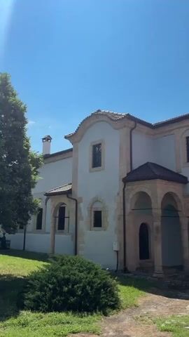 The video shows a side view of a light blue building with a light brown roof, featuring a colonnade with arched openings. The building has several windows, some of which are dark with visible grills. In front of the building, there is green grass and a dark green bush. The sky is clear and blue with no clouds. The video pans slightly to the right, revealing more of the building's facade and the surrounding trees.