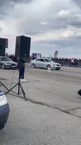 The video captures two BMW cars performing a burnout in front of a large crowd. A silver BMW sedan is on the right side of the screen, and a gray BMW coupe is on the left. Both cars emit a cloud of smoke as they accelerate. In the foreground, a photographer kneels, capturing the scene. A person in a black tracksuit with the letters "VTA SECURITY" stands on the right edge of the screen, facing towards the cars. The sky is overcast with dark clouds. A black stage or wall is visible to the left, along with a green flag and a sign that reads "7." A metal fence separates the crowd from the racing area. The background reveals a field under the cloudy sky.