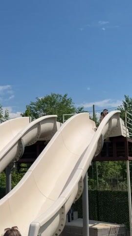 A person is going down a water slide. A second person is sitting on the slide waiting their turn. A third person is already in the pool at the bottom of the slide. The water slide is made of white plastic and is supported by metal poles. The water slide is surrounded by trees and a blue sky.