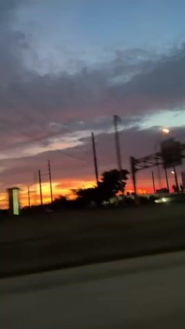 This is a blurry, vertical video of a sunset viewed from a moving vehicle. The sky is filled with streaks of orange, pink, and purple clouds. In the foreground, there is a blurry highway with concrete barriers. In the middle ground, there are silhouetted objects including what appears to be a traffic light or sign structure and some buildings. The motion blur suggests the camera is moving at a considerable speed.