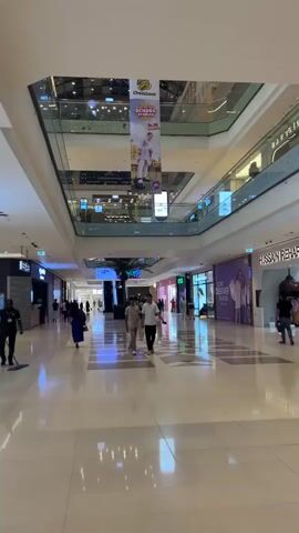 A busy shopping mall interior is depicted. The camera pans from left to right, showing a wide corridor with shops lining both sides. Several people are walking through the mall. On the left side, a security guard is mopping the floor. To the right of the security guard, a family walks by, including two adults and two children. Further down the corridor, a woman in a blue dress walks away from the camera. Two men in light-colored clothing walk towards the camera. Overhead, banners are displayed advertising a "School by Cheezious" promotion. The mall features multiple levels, with escalators visible in the distance. The lighting is bright, and the floor is highly reflective.