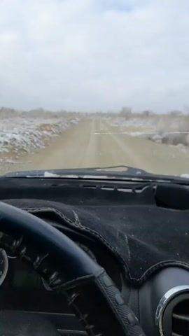 The video shows a car's dashboard and steering wheel from the driver's perspective, traveling on a dirt road. Patches of snow are visible on either side of the road. The sky is overcast.