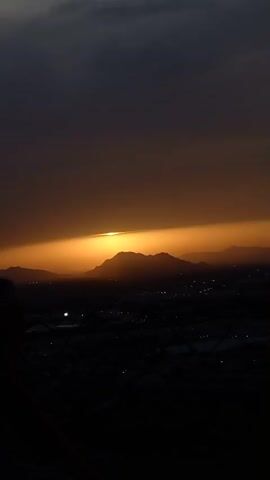A silhouetted mountain range is visible against a dark, cloudy sky with an orange glow emanating from the setting sun, which is partially obscured by the mountains. Below the mountains, a dark landscape is dotted with faint lights, suggesting a town or city. The overall scene captures the dramatic transition from daylight to night.