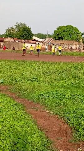 A group of young men in yellow jerseys are playing soccer on a dirt field. There are spectators watching the game from the side of the field. The field is surrounded by a few small buildings with corrugated metal roofs. The area is overgrown with grass and vegetation. The sky is clear and the sun is shining.