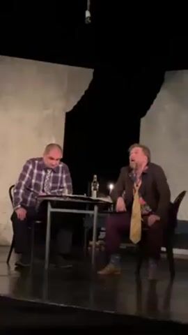Two middle-aged men are seated at a small table on a stage. One is wearing a brown suit with a yellow tie, a floral shirt, and patterned socks. The other is wearing a purple and white checkered shirt with a tie, and dark pants. A bottle of alcohol sits on the table between them. They both appear to be looking upwards. They are on a stage with a backdrop that suggests a wall. The lighting is dim.