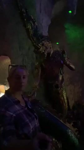 A blonde woman wearing a plaid shirt and glasses stands in the foreground of a dimly lit cave-like environment. Behind her, a large, elaborate dragon sculpture is visible, adorned with intricate details and illuminated by green and blue lights. The woman appears to be looking towards the camera with a neutral expression. The overall atmosphere suggests an indoor attraction or display, possibly a themed exhibit.