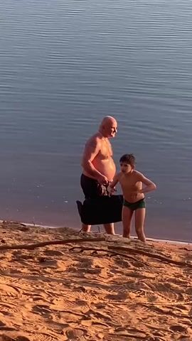 A man and a boy stand at the edge of a body of water, possibly a lake or the ocean, at what appears to be a sandy beach. The boy, wearing green swimming trunks, stands in the shallow water near the shore. He is looking off to the side, and his posture suggests he might be cold. Behind him, a shirtless, bald man stands on the beach. He is wearing dark shorts and carrying a black bag. The sand appears to be reddish or golden in color. The water is calm with subtle ripples, reflecting a muted blue or grey light. There are a few fallen twigs and branches scattered on the sand. The lighting suggests it might be late afternoon or early morning.