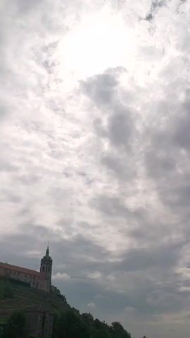 The video shows a low-angle shot of a cloudy sky above a building with a tower. The building appears to be on a hill, and there are trees in the foreground. The sky is mostly gray with some brighter patches, suggesting the sun is behind the clouds. The clouds are moving, creating a sense of depth and movement in the sky.