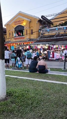 The camera captures a bustling scene at Hoi An Market in Vietnam. In the foreground, two people are seated on a grassy patch, taking a moment to rest.  Beyond them, a lively street market is filled with vendors displaying an array of colorful clothing and souvenirs.  Numerous people browse the stalls, creating a dynamic and energetic atmosphere.  The market is housed in a bright yellow building with the name "CHO HOI AN MARKET" prominently displayed above the entrance.  Red lanterns hang, adding a festive touch. The overall scene is a vibrant snapshot of Hoi An Market, showcasing its charm and popularity among visitors.