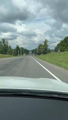 A vertical, first-person perspective video captures a view from inside a vehicle as it travels down a rural asphalt road. The road is lined with trees on both sides and stretches towards a distant horizon under a cloudy sky. Several vehicles are visible on the road ahead. A yellow sign with a blue border and text is posted on the right side of the road. The lower portion of the video shows the car's dashboard and windshield wipers. The road has white lane markings. A road sign depicting a child running is painted on the asphalt in the foreground.