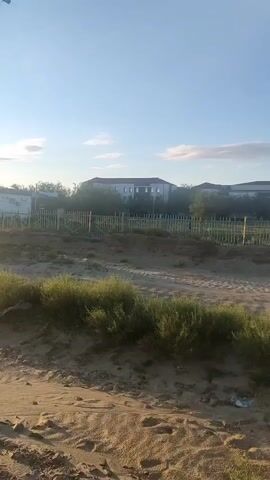 A video captures an outdoor scene on a clear, sunny day. The camera pans across a landscape featuring sandy terrain with sparse vegetation. In the background, several buildings are visible, one appearing to be a multi-story structure. A metal fence runs along a line of trees, separating the foreground from the background structures. A utility pole with wires stands prominently in the middle ground. The sun is bright, casting shadows and creating lens flare in some shots. The overall environment appears to be somewhat arid or undeveloped.