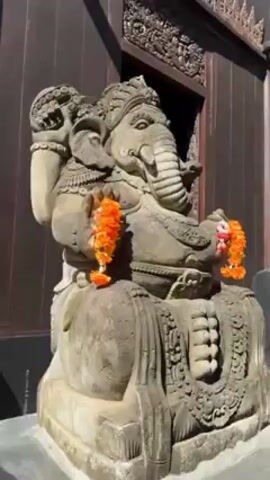 The video shows a close-up of a stone Ganesha statue decorated with orange and white garlands, followed by a view of a dark wooden building with intricate carvings. The camera then pans up to reveal the pointed roof of the building against a clear blue sky and trees. Finally, the camera shows the entire building, revealing the ornate entrance and a man standing in front. The building appears to be a temple or cultural site with traditional Southeast Asian architecture.