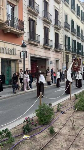 The video shows a street scene with a religious procession. The procession consists of people wearing brown robes and tall, pointed white hoods. One person carries a large wooden cross, while another carries a banner with a religious image. People are standing on the sidewalk watching the procession. The buildings lining the street are multi-story and feature balconies and shutters. The sky is overcast. A small garden bed with plants and irrigation lines runs along the edge of the street.