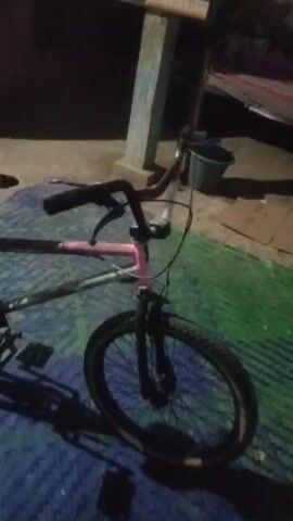 The video shows a BMX bicycle at night. The bicycle is on a blue mat and there are shadows cast on the ground. The bicycle is red and pink, with black handlebars, a black seat, and black tires. The video pans around the bicycle, showing the front wheel, the handlebars, the frame, the seat, the rear wheel, and the pedals.
