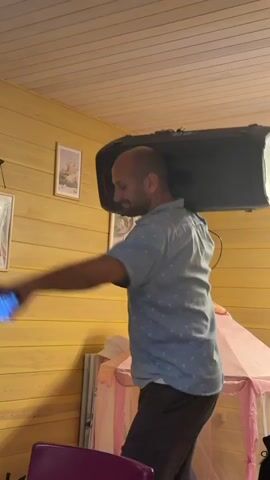 A bald man with a beard, wearing a light blue short-sleeved shirt and dark pants, carries a large black speaker on his right shoulder. He is indoors, in a room with yellow wood-paneled walls and a wood-paneled ceiling. There are two framed pictures on the wall behind him. To his right, a pink children's tent is visible. The man is smiling and appears to be dancing or moving rhythmically. His left hand is extended forward, and he is holding a dark object, possibly a phone. A tripod with a phone is also visible in the lower left corner of the frame. The lighting is dim.