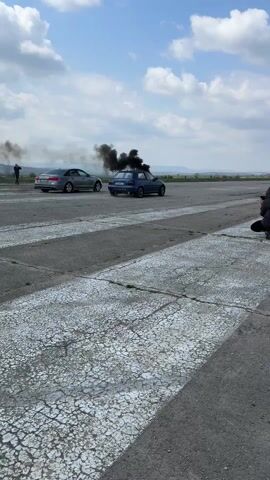 The video shows two cars doing burnouts on an abandoned airstrip. Both cars are emitting a lot of black smoke. A photographer in a black hooded jacket is squatting on the runway, filming the cars as they burn out. A person is standing on the left of the screen watching the cars burn out as well. The runway is gray and cracked with white lane markers. The sky is blue with some white clouds. The horizon shows a field in the background.