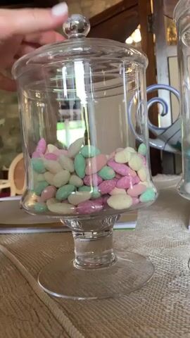 The video shows a close-up of a clear glass jar filled with pastel-colored candies. The jar has a pedestal base and a handle with a blue and white swirl design. The candies are oval-shaped and appear to be a mix of pink, light green, and cream colors. A person's hand, with a white manicure, is seen reaching into the jar to pick up a candy. The background suggests an indoor setting with stone walls and wooden furniture, hinting at a rustic or historical location. The lighting appears to be natural, possibly from a window, indicating daytime. The overall mood is casual and perhaps suggests a moment of indulgence or preparation for an event. The location is identified as Castiglione d'Orcia, Italy. There is no visible text in the media.