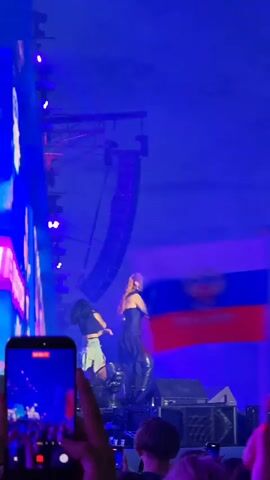 The video shows a concert with two female singers on stage. The singer on the right has long red hair and is wearing a black dress. She is holding a microphone and singing. The singer on the left has dark hair and is wearing a crop top and ripped jeans. She is also holding a microphone and singing. They are both standing on a stage with a large screen behind them. There are stage lights and speakers on the stage. The audience is visible in the foreground, holding up phones to record the performance. Some audience members are also waving Russian flags. The overall atmosphere is of a lively music event.