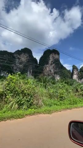 The video shows a car driving on a dirt road past a lush green landscape dominated by towering limestone karsts. The karsts are jagged and covered in dense vegetation. The sky is blue with scattered white clouds. There are power lines strung across the frame. On the right side of the road, there is a white sign with directional arrows and text in a foreign language, likely indicating local destinations. The car's side mirror is visible in the lower right corner of the frame. The camera appears to be mounted inside the vehicle, looking out the passenger side window.