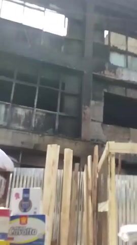 The camera pans up to reveal a derelict building with charred remains, indicating a possible fire. The building appears to be two stories tall, with broken windows and significant structural damage. There is a makeshift fence constructed from wooden planks and corrugated metal in front of the building. Some objects, potentially merchandise, are visible under a white sheet near the fence. The overall scene conveys a sense of abandonment and destruction.